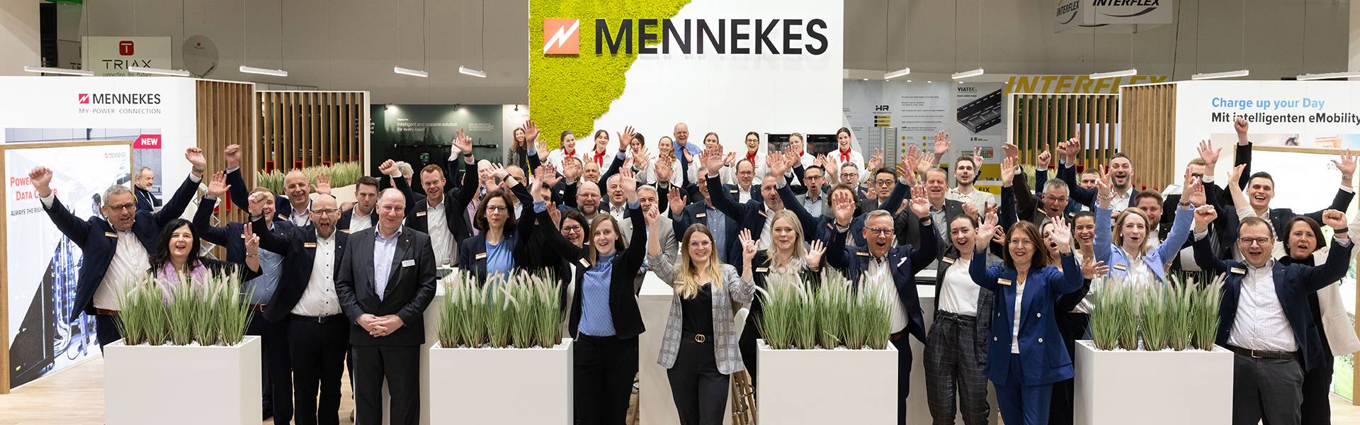 Mitarbeitende von MENNEKES stehen auf einem Messestand und heben jubelnd die Hände, im Hintergrund das MENNEKES-Logo.
